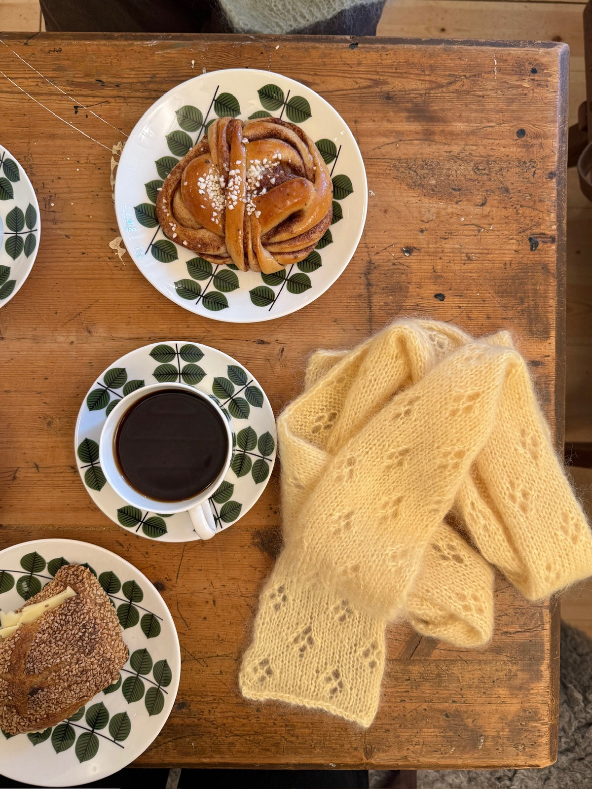 Coffee, a cinnamon bun and a pale yellow scarf on a wooden table